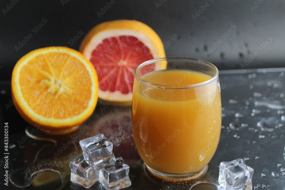 The juice of orange abicos yellow in the glass next to lie half of orange and grapefruit ice on a black background. Refreshing Drinks Summer The juice of orange abicos yellow in the glass next to lie half of orange and grapefruit ice on a black background. Refreshing Drinks Summer