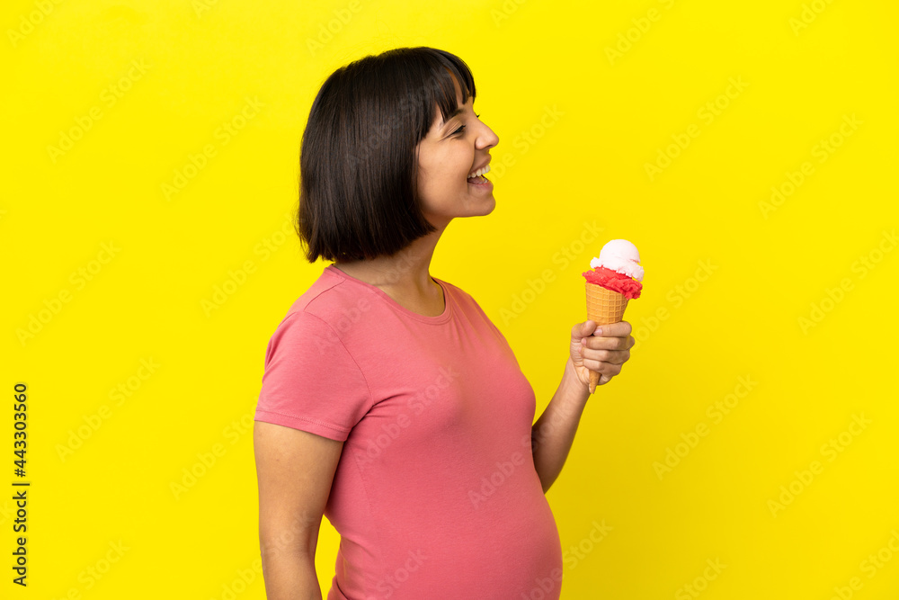 Pregnant woman holding a cornet ice cream isolated on yellow background laughing in lateral position Pregnant woman holding a cornet ice cream isolated on yellow background laughing in lateral position