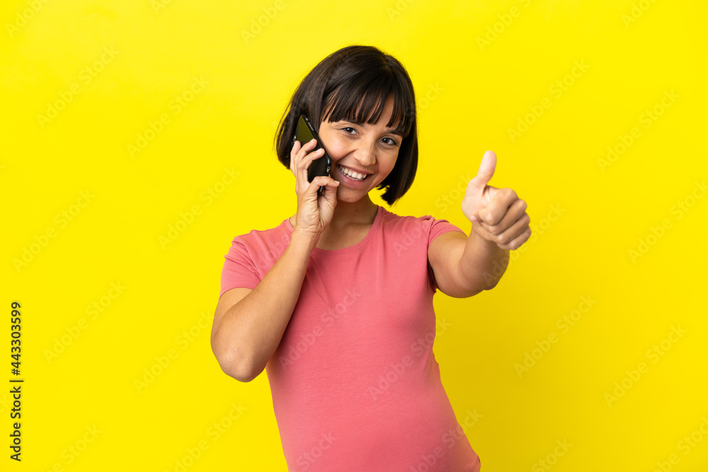 Young mixed race pregnant woman isolated on yellow background keeping a conversation with the mobile while doing thumbs up Young mixed race pregnant woman isolated on yellow background keeping a conversation with the mobile while doing thumbs up