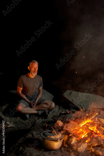 Peaceful mature man, tourist sitting with eyes closed near campfire and cooking dinner outdoors in forest at night