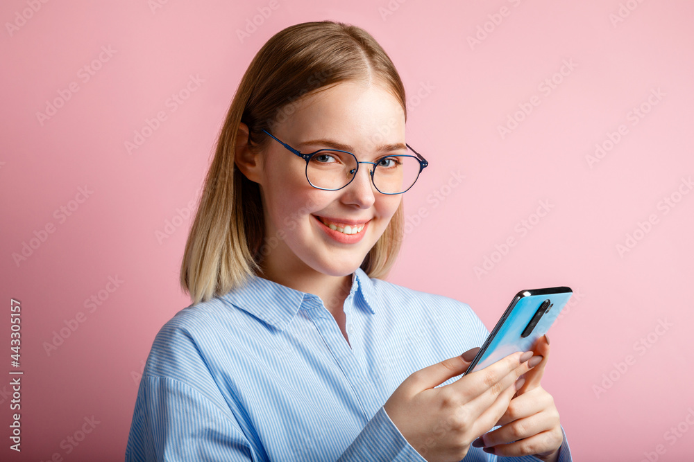 Emotional portrait of happy positive smile manager office worker in glasses use smartphone. Woman in a blue shirtwrites a message on phone isolated over color pink background.