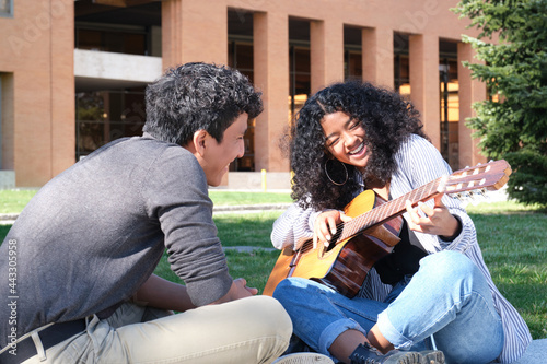 Young latin man teaching how to play the guitar to a young latin woman laughing at the university campus. University life, millennial generation.