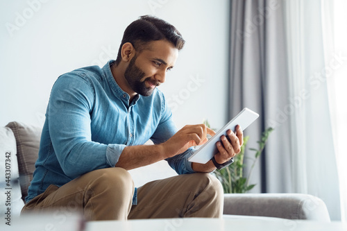 Millennial indian man using tablet reading news browsing internet, watching videos, checking social networks relaxing on couch sofa in living room. Online work, education or communication concept