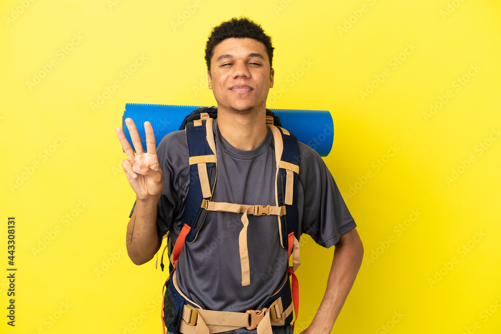 Young mountaineer African American man with a big backpack isolated on yellow background happy and counting three with fingers Young mountaineer African American man with a big backpack isolated on yellow background happy and counting three with fingers