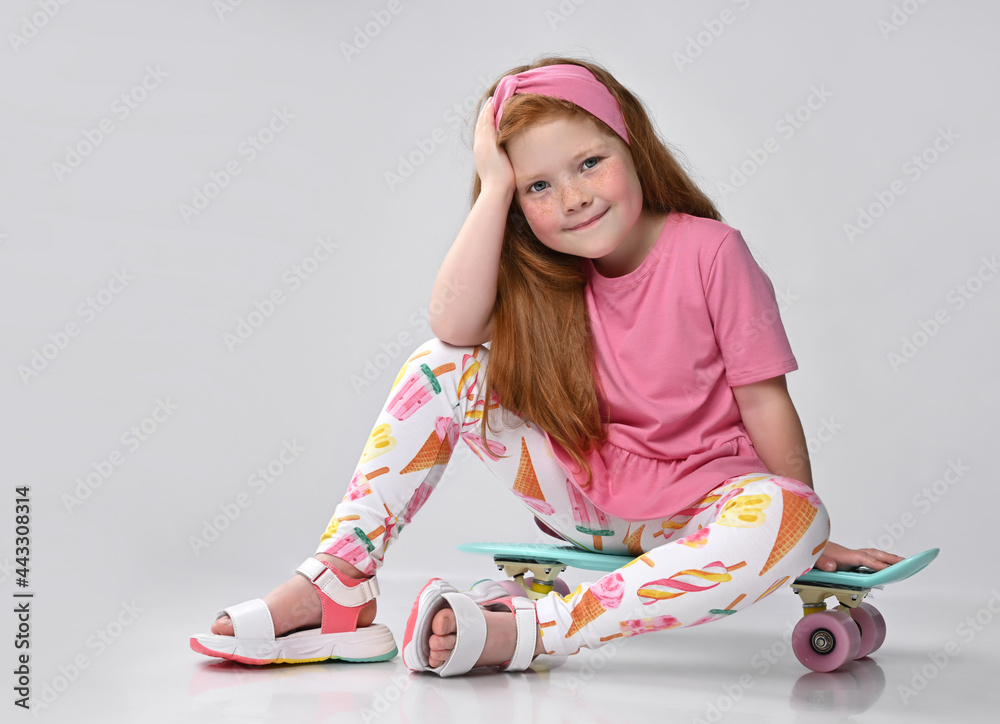Smiling red-haired little girl in a summer dress fashion, sitting on a skateboard, and props up a head a hand, gazing at the camera with satisfaction and happiness. Positive emotions of children