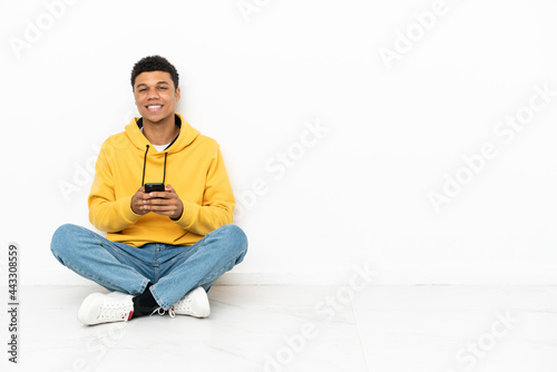 Young African American man sitting on the floor isolated on white background sending a message with the mobile