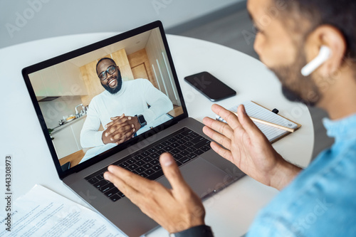 Over shoulder view Indian man sitting at home desk front of laptop greeting business partner on screen. Freelance entrepreneur businessman communicate remotely online with colleague using pc computer photo