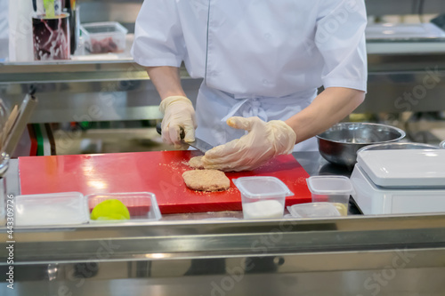 Chef hands holding knife and preparing breaded minced meat cutlet on red cutting board - close up. Professional cooking, cookery, gastronomy and food concept