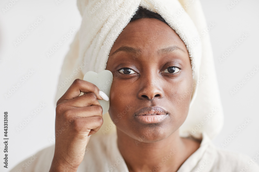 Front view portrait of beautiful African-American woman doing face massage while enjoying skincare routine at home and looking at camera, copy space Front view portrait of beautiful African-American woman doing face massage while enjoying skincare routine at home and looking at camera, copy space
