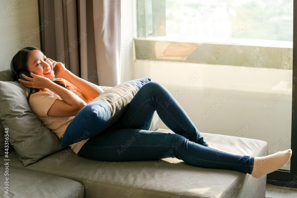 Beautiful Asian woman lying down on sofa by the window in apartment and listening to the music on headphone with smiling face. Female enjoy indoor lifestyle feel relax and comfortable resting on couch Beautiful Asian woman lying down on sofa by the window in apartment and listening to the music on headphone with smiling face. Female enjoy indoor lifestyle feel relax and comfortable resting on couch
