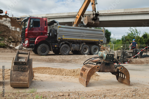 Close-up shot of the disassembled hyraulic pliers and bucket for the excavator lying on the ground at the demolition site. In the background the excavator is moving the debris into the truck.