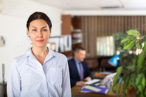 Closeup portrait of positive confident adult Asian woman wearing light blue shirt standing in modern office, selective focus