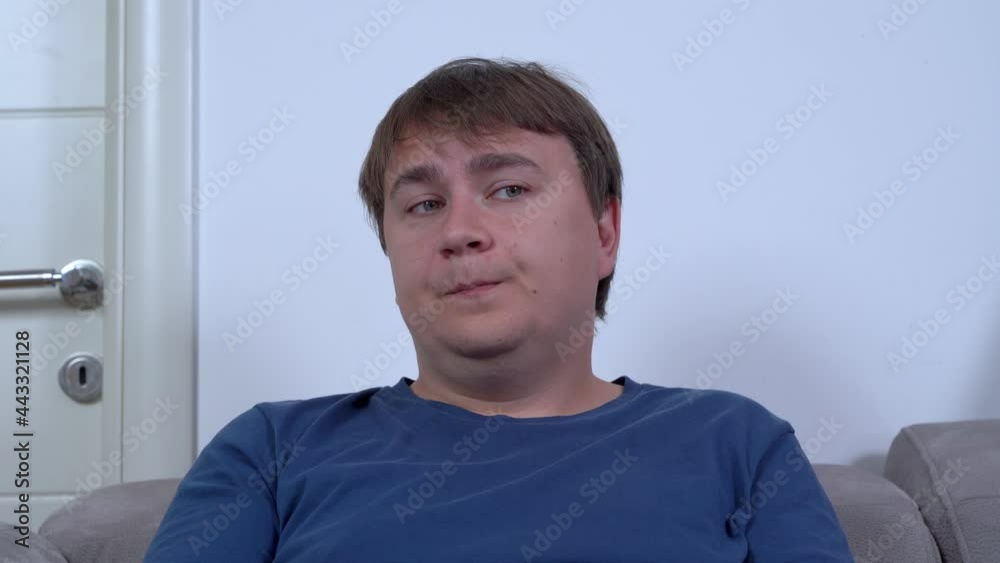 Crafty plump man wearing blue t-shirt shrugs letting eyes rove and grimacing on comfortable grey sofa in living room closeup Crafty plump man wearing blue t-shirt shrugs letting eyes rove and grimacing on comfortable grey sofa in living room closeup