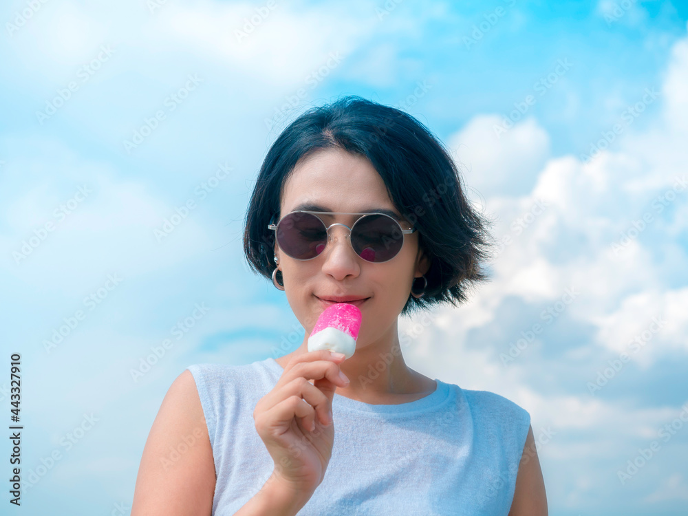 Smiling woman eating popsicles. Happy beautiful Asian woman short hair wearing sunglasses and casual white sleeveless shirt holding pink popsicle on blue sky background in summer.