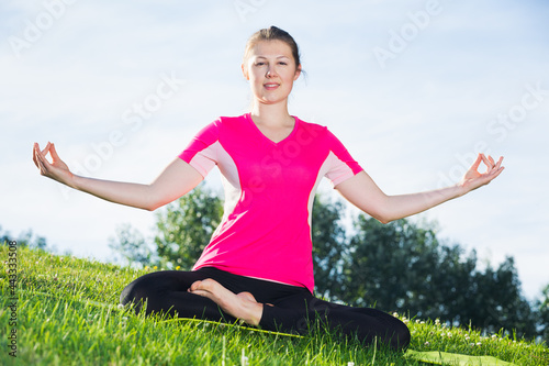 Female 20-30 years old in pink T-shirt is sitting and meditating in the park.