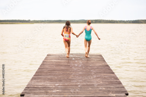 2 girls on holiday running on wooden bridge to jump into water  rear view  summer time  vacations