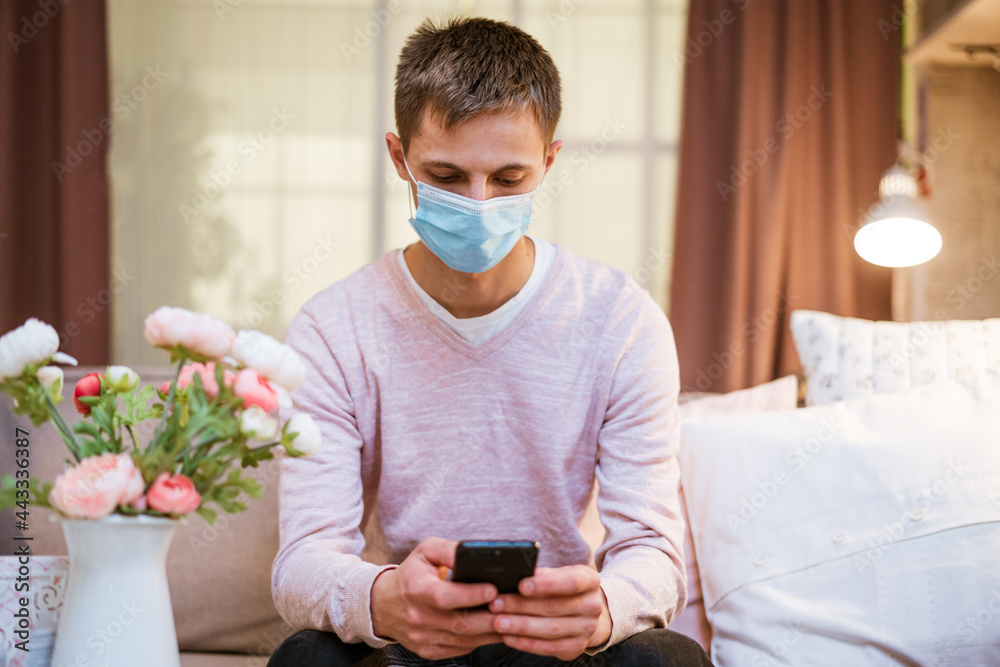 young caucasian man sitting on a sofa in casual clothes, wearing a protective medical mask with a phone in his hand