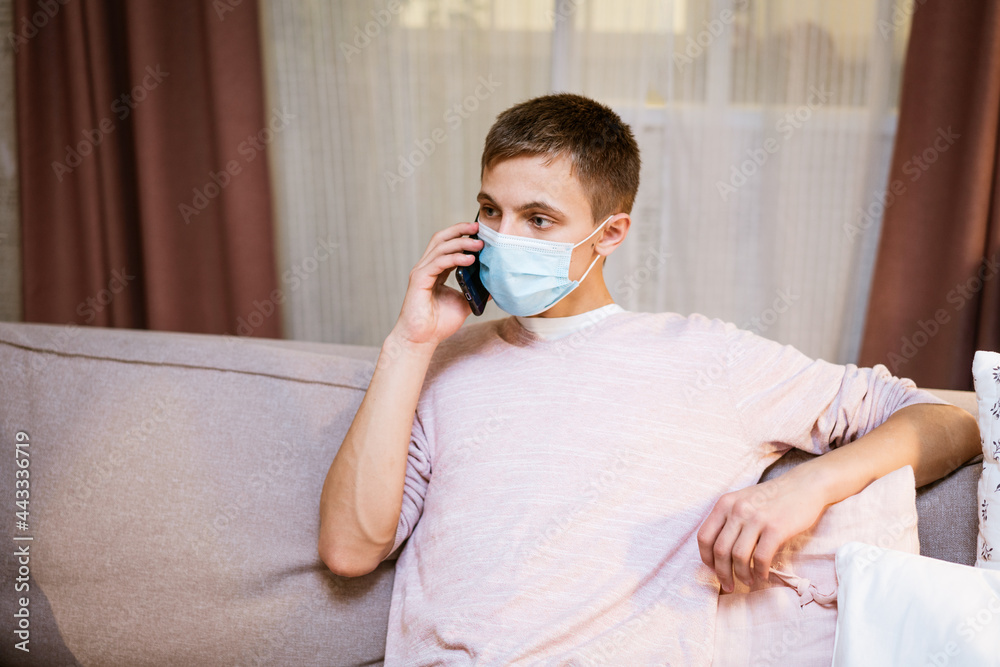 a young man of Caucasian appearance, in casual clothes, talking on the phone in a protective mask while sitting on the couch at home. The concept of remote communication
