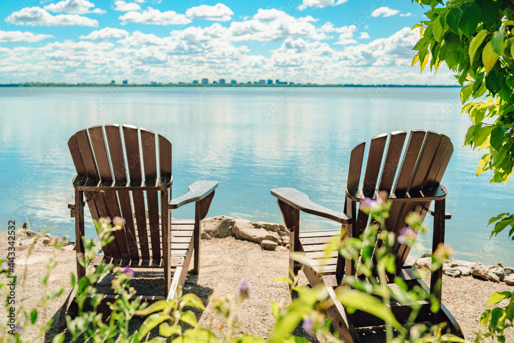 Fototapeta premium Two Muskoka chairs by the water on home terrace with calm view of lake in Canada. Summer cottage vacation lifestyle.