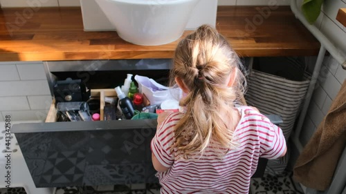 Fair haired preschooler girl in striped t-shirt opens large bathroom drawer and looks inside with interest searching tools at home