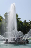 Tall fountain in the park inside a pool of water in front of green trees and blue sky