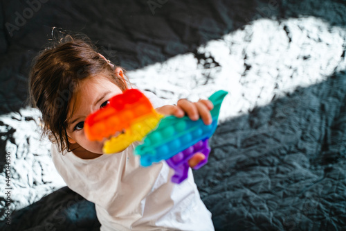 Toddler girl with popit toy in rainbow colours. Dino shape silicone toy for stress relief. Bubbles sensory trendy fidget. Child playing at home in candid style