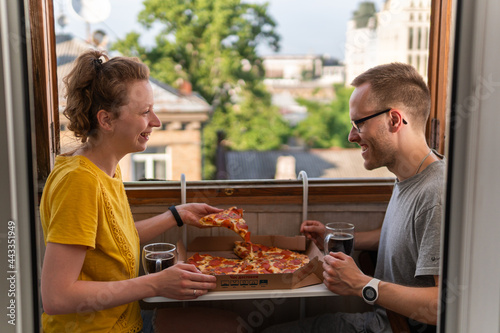 A young happy couple is having meal  dinner  enjoying pizza  salad and drinking Kvass at home on the balcony with open windows  amazing view and portable compact table