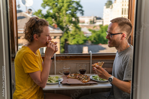 A young happy couple is having meal, dinner, enjoying pizza, salad and drinking Kvass at home on the balcony with open windows, amazing view and portable compact table