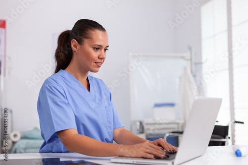 Medical nurse in uniform using laptop sitting at desk in hospital office. Health care physician using computer in modern clinic looking at monitor, medicine, profession, scrubs.