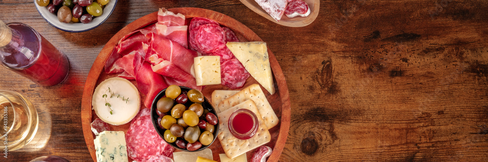 Charcuterie and cheese platter with wine and olives panorama, shot from above on a dark rustic wooden background with a place for text. Italian antipasti or Spanish tapas panoramic banner