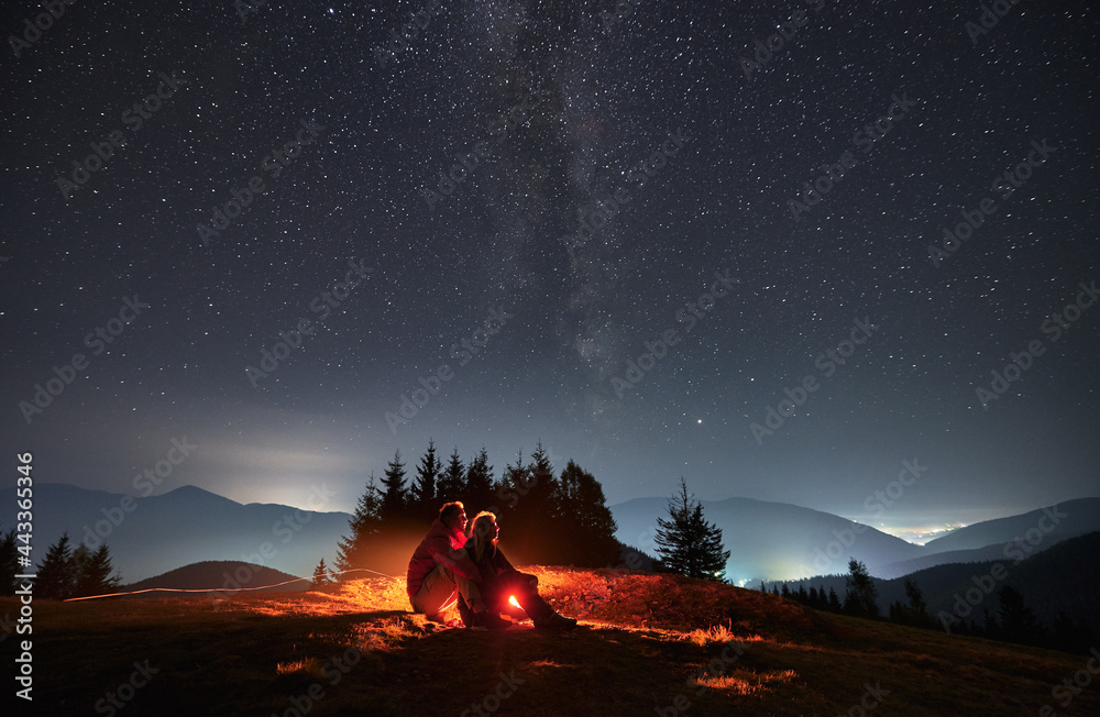 Young man and woman travelers sitting on grassy hill under night sky with stars. Couple hikers looking at majestic night starry sky and hugging. Concept of hiking, night camping and relationships. Young man and woman travelers sitting on grassy hill under night sky with stars. Couple hikers looking at majestic night starry sky and hugging. Concept of hiking, night camping and relationships.
