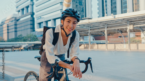 Asian businesswoman go to work at office stand and smiling wear backpack look at camera with bicycle on street around building on a city. Bike commuting, Commute on bike, Business commuter concept. photo