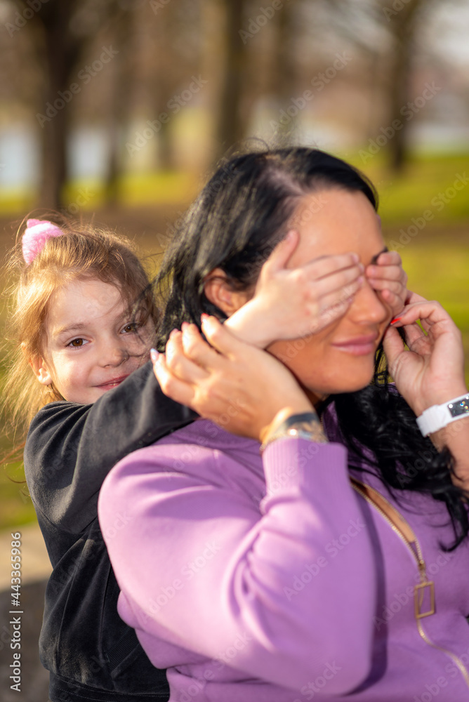 A young little girl covered her mother's eyes.Positive young woman smiling at summer evening park.Closeup. A young little girl covered her mother's eyes.Positive young woman smiling at summer evening park.Closeup.