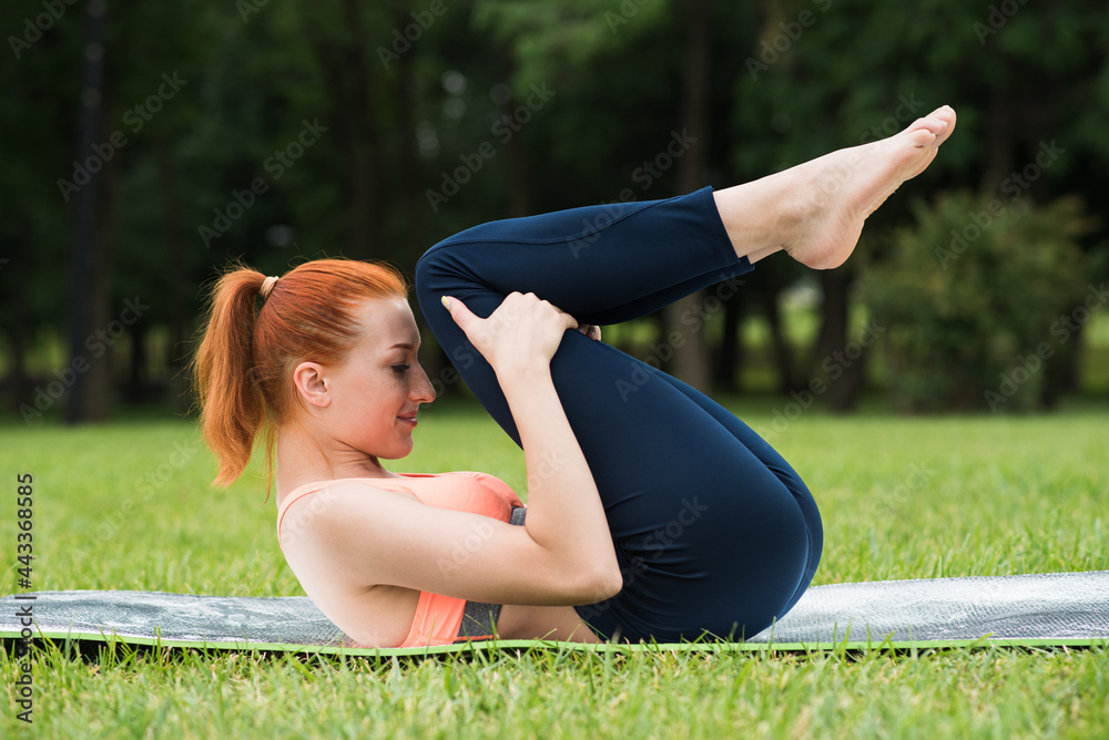 Fototapeta premium Attractive redhead girl practicing yoga while lying on a mat on the grass in the park