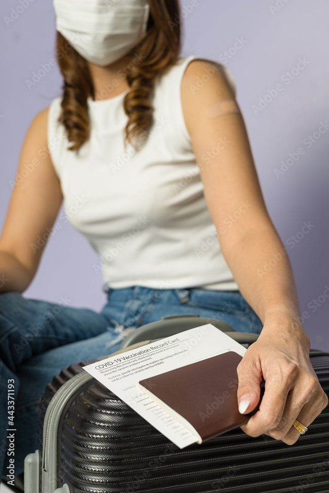 Fototapeta premium Vaccinated woman wearing hygiene protective medical mask holding Covid-19 coronavirus vaccination record card and passport on travel bag. The concept for life after a virus outbreak. New normal idea