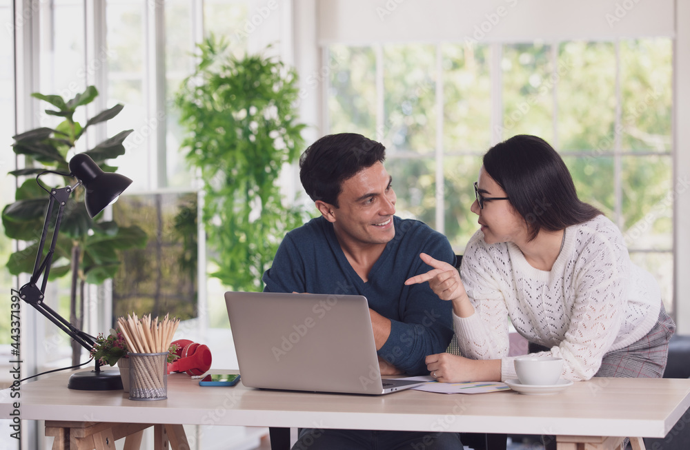 Caucasian man and Asian woman mix race lovers working together via laptop notebook computer and discussing in living room with happy and intimate. New normal work at home office concept Caucasian man and Asian woman mix race lovers working together via laptop notebook computer and discussing in living room with happy and intimate. New normal work at home office concept