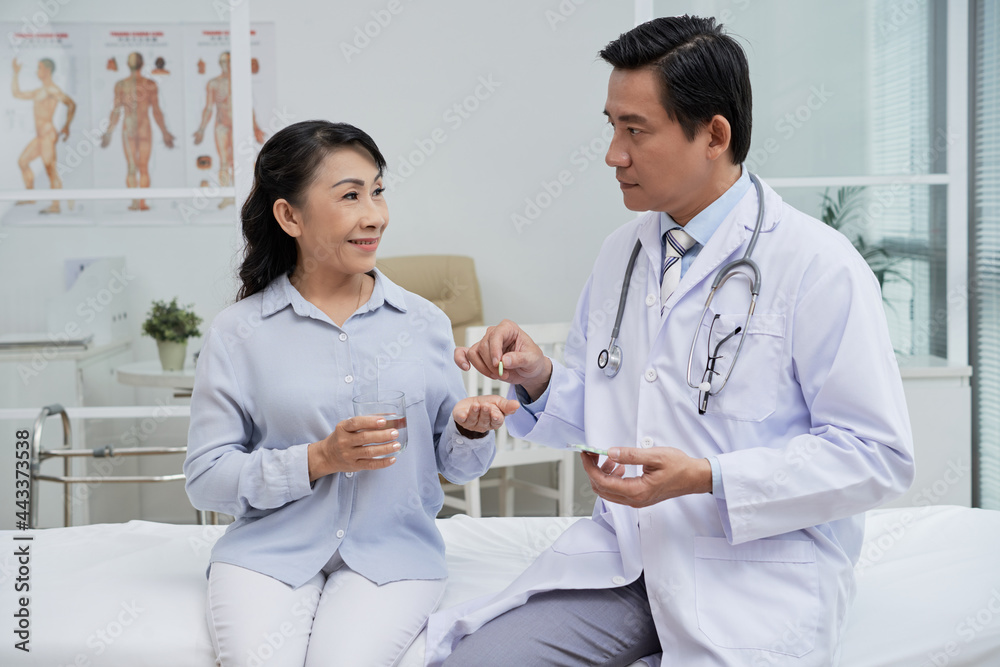 Serious physician giving pills to smiling senior woman sitting on bed with glass of water Serious physician giving pills to smiling senior woman sitting on bed with glass of water