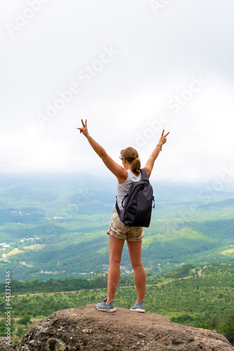 Happy woman with backpack standing on edge of cliff and looking at valley with her hands raised. Summer landscape with sporty girl, green grass, forest, hills , blue sky in fall