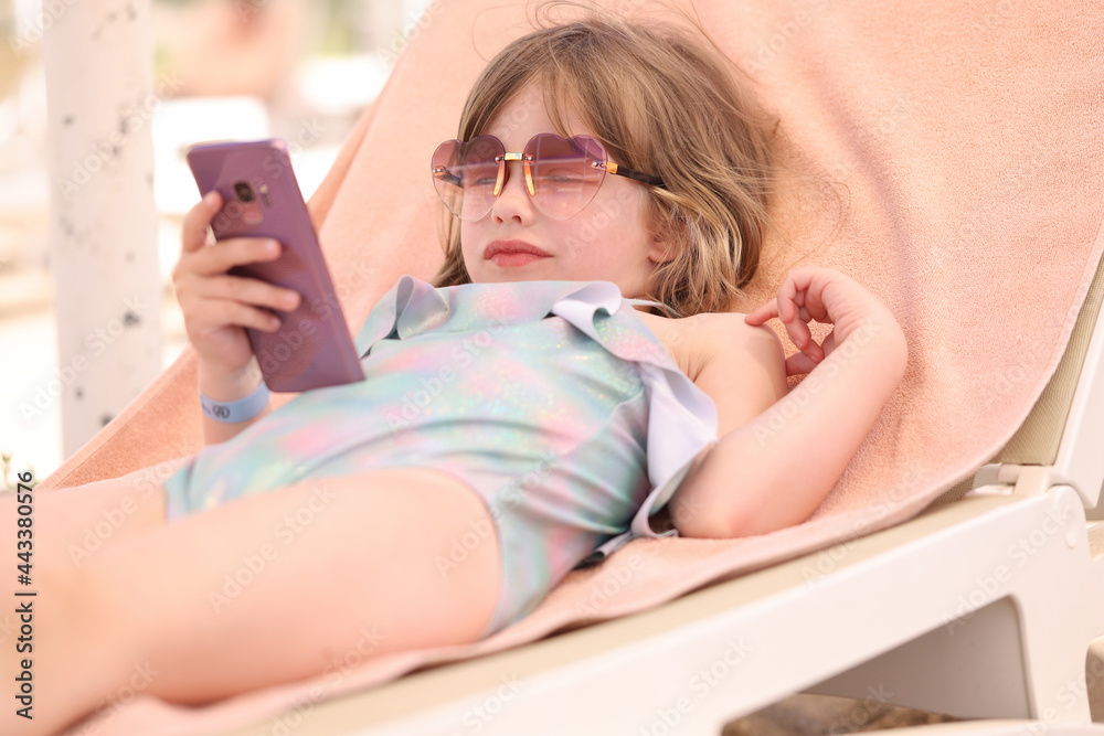 Little girl in sunglasses and swimsuit lying on lounger with mobile phone in her hands Little girl in sunglasses and swimsuit lying on lounger with mobile phone in her hands