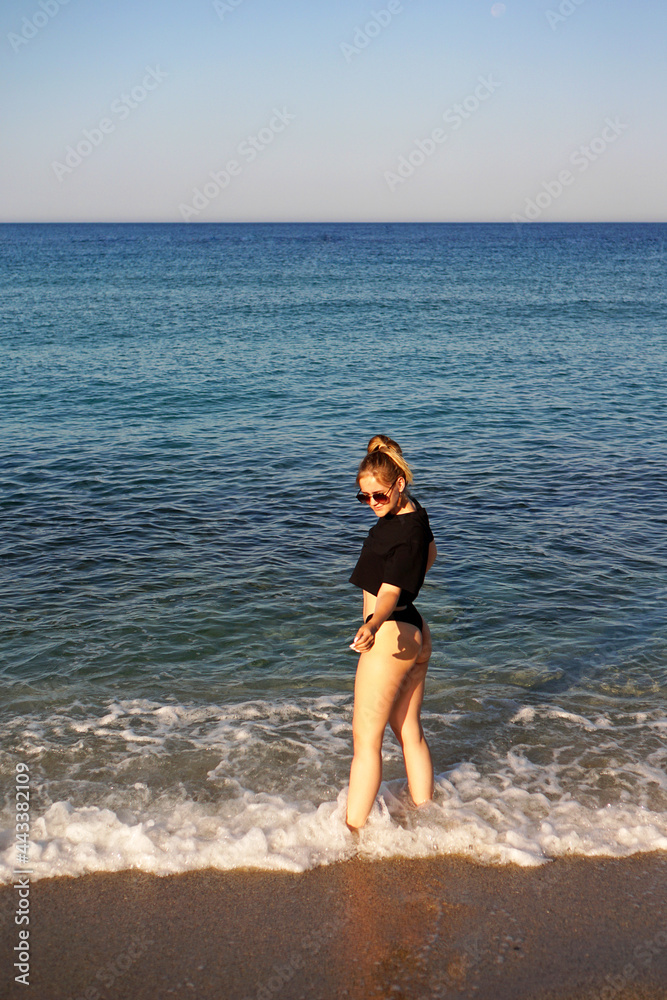 Fototapeta premium Young woman in a black tank top and underpants on the sea beach. Tanned body, body positive. Vertical photo