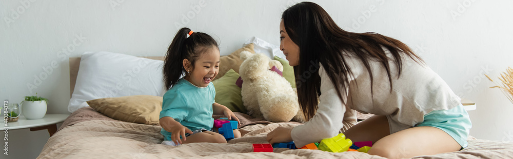 Asian mother and excited kid playing building blocks on bed, banner Asian mother and excited kid playing building blocks on bed, banner
