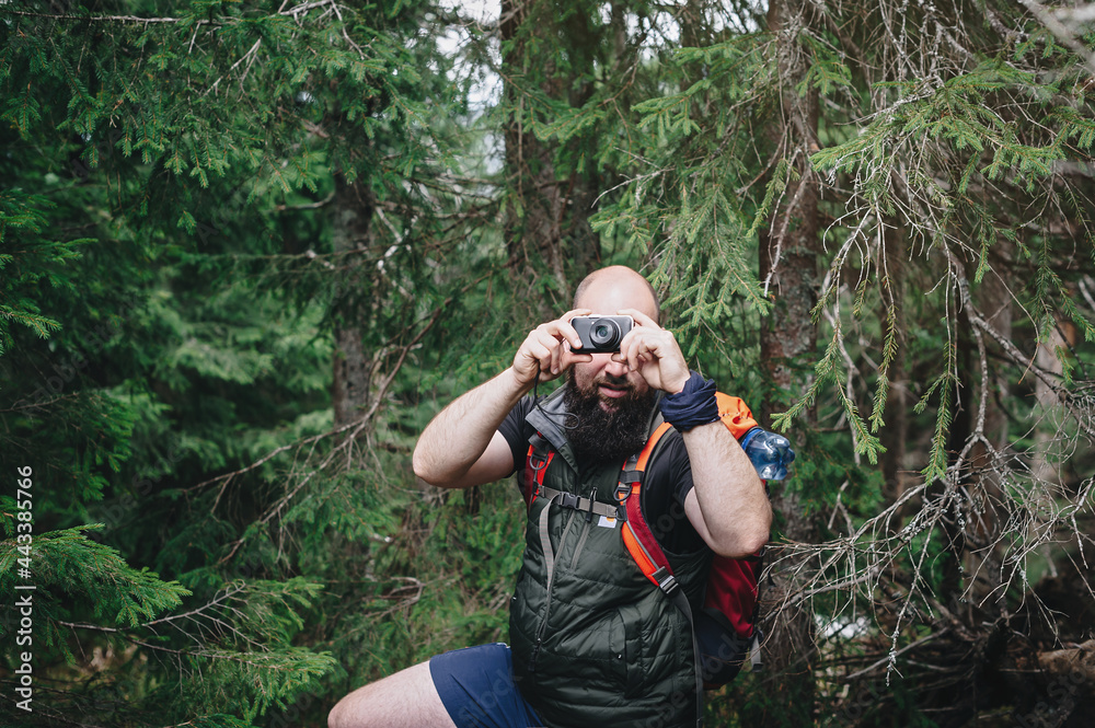 A young hipster man with a backpack on his back stopped to photograph a beautiful landscape in the woods on a camera Tourist on a hike takes photos on digital camera. A young hipster man with a backpack on his back stopped to photograph a beautiful landscape in the woods on a camera Tourist on a hike takes photos on digital camera.