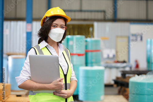 woman engineering wear protective face mask and safety helmet holding laptop standing in warehouse factory. manufacture production of hygiene masks industry. photo