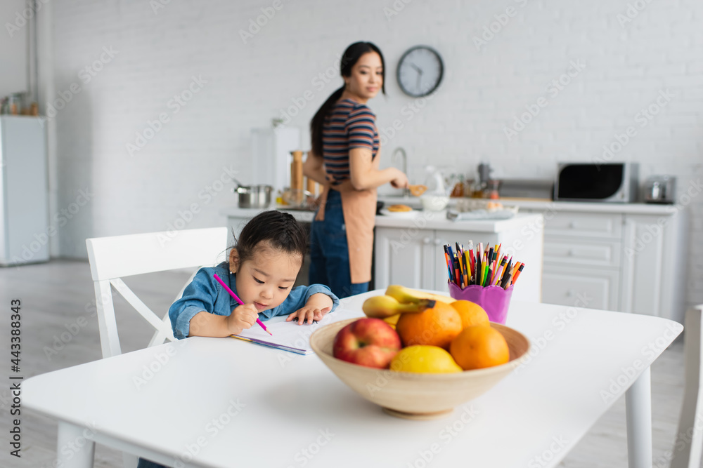 Asian kid drawing near fruits and blurred mother at home