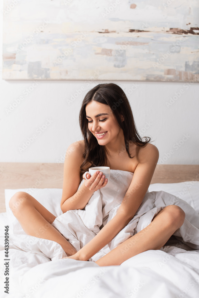 young brunette woman smiling while holding cup of coffee in bed young brunette woman smiling while holding cup of coffee in bed