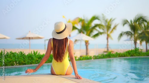 Backside view of the woman sitting on edge of swimming pool at exotic hotel in Hawaii near the beach in yellow monokini and white hat and hitting water with her legs, beach umbrellas and Palms photo