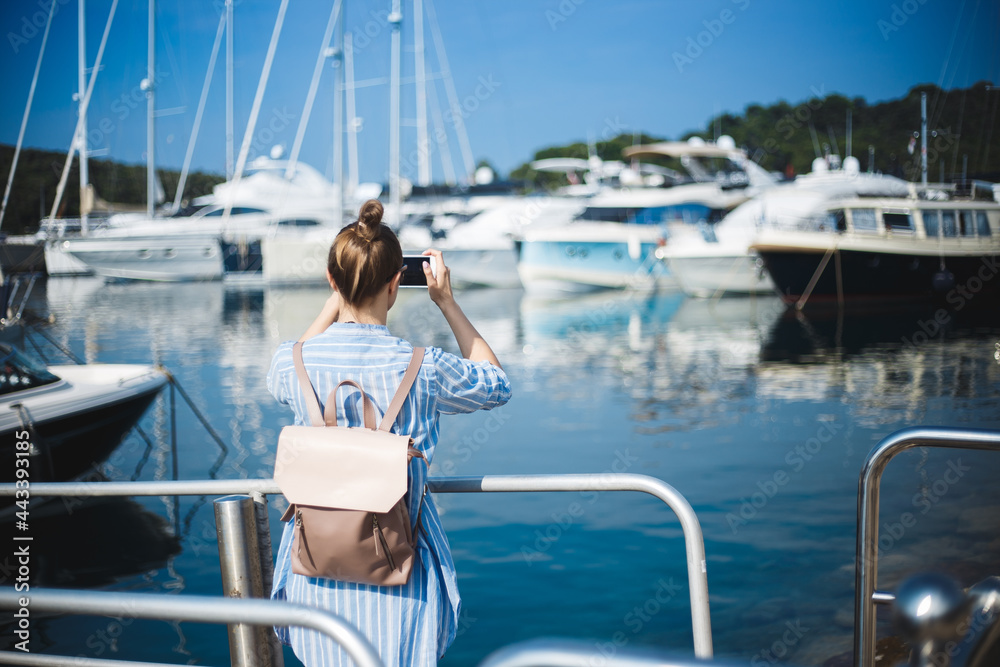 A girl-blogger is photographing yachts on a smartphone. White Caucasian girl in blue dress and stylish backpack on the background of blue sea and yachts.