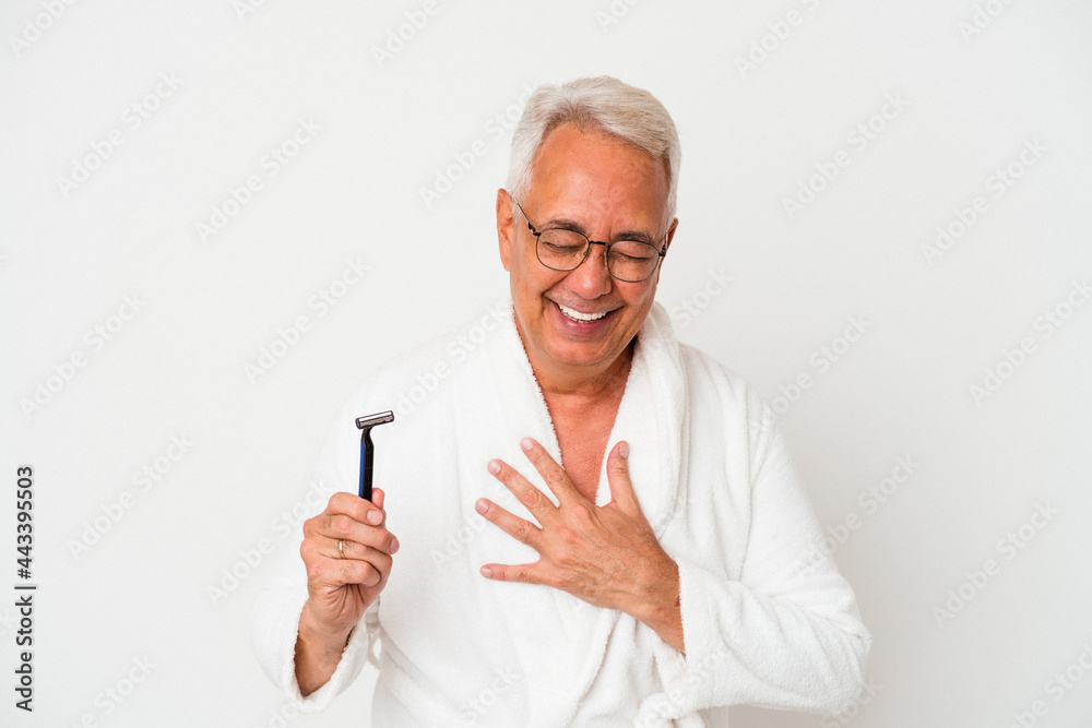 Senior american man wearing bathrobe holding razor blade isolated on white background laughs out loudly keeping hand on chest. Senior american man wearing bathrobe holding razor blade isolated on white background laughs out loudly keeping hand on chest.