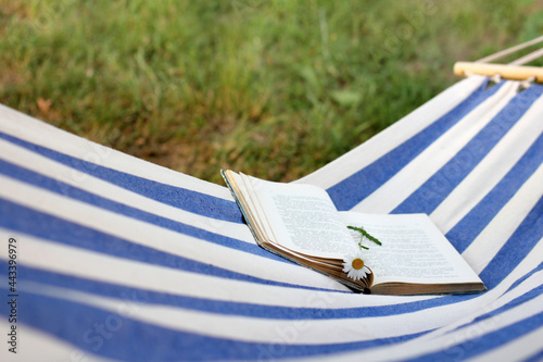 open book with a bookmark of a chamomile flower on a hanging hammock in the garden. outdoor reading in summer