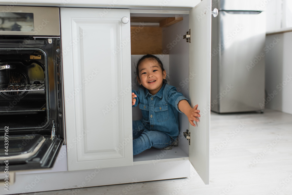 Smiling asian girl looking at camera from kitchen cabinet Smiling asian girl looking at camera from kitchen cabinet